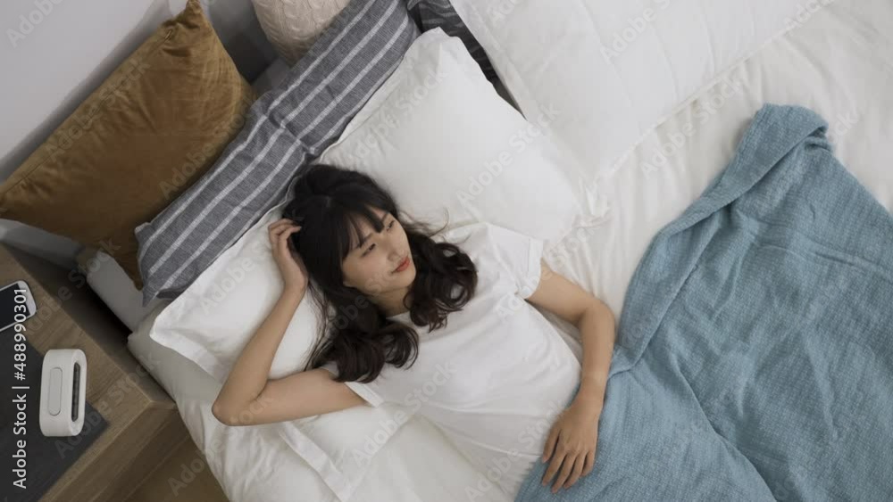 zoom out with view from above smiling asian girl lying awake in bed is gazing into the distance and enjoying tranquil morning with hand above her head at home. zoom out with view from above smiling asian girl lying awake in bed is gazing into the distance and enjoying tranquil morning with hand above her head at home.