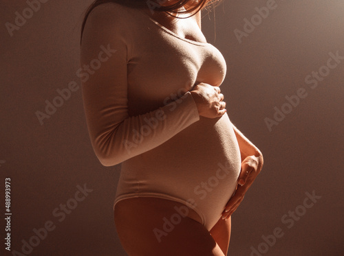 pregnant body. pregnant girl with red hair is standing in warm light in studio with hands on pregnant stomach on wall background. pregnant concept, free space