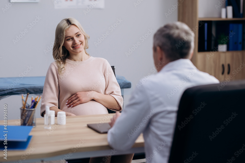 Obraz premium Smiling pregnant woman talking to male doctor at hospital during pregnancy consultation. Happy expecting mother visiting gynecologist.