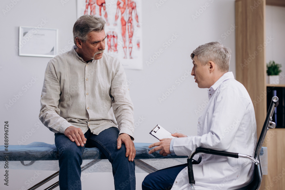 Obraz premium Grey haired man patient talking to male GP doctor. Pensioner complaining about knee pain, explaining his health condition.