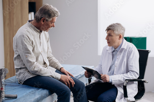 Grey haired man patient talking to male GP doctor. Pensioner complaining about knee pain, explaining his health condition.