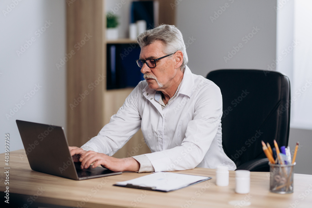 Serious male doctor sitting at workplace in hospital working on computer, looking thoughtful. Professional physician studying patient's electronic medical record. Serious male doctor sitting at workplace in hospital working on computer, looking thoughtful. Professional physician studying patient's electronic medical record.