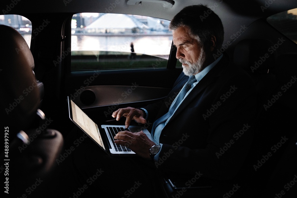 Full concentration at work. Confident mature man in full suit working using laptop while sitting in the car.