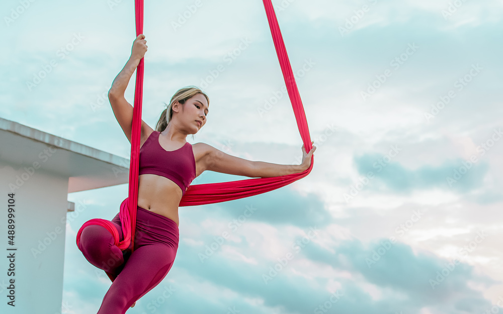 Beautiful healthy strong woman wearing sport bra, relaxing, exercise yoga fly in air, aerial hoop on outdoor rooftop in morning. There are background of sky, sunlight flare, copy space advertisement.
