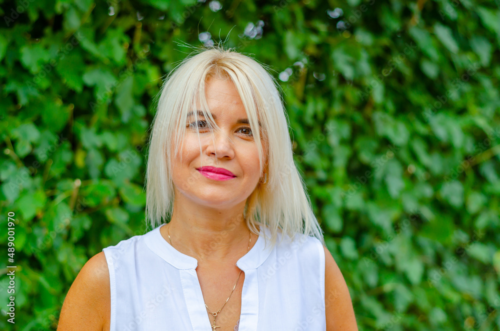 Portrait of a tanned middle-aged woman. Bright makeup. Blonde. Tousled white hair. Blurred greens background. Portrait of a tanned middle-aged woman. Bright makeup. Blonde. Tousled white hair. Blurred greens background.