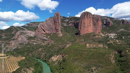 Clear blue river meanders in the valley while in the background the high orange steep mountain walls of Mallos de Riglos stand prominently in the Spanish nature. Wide drone dolly shot photo