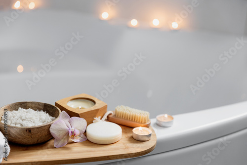 Preparation for hotel spa treatment, home bath procedure. White washbasin in bathroom, accessories on tray. Burning candles, soap, foot brush, towel, glass bottle with sea salt, orchid flower