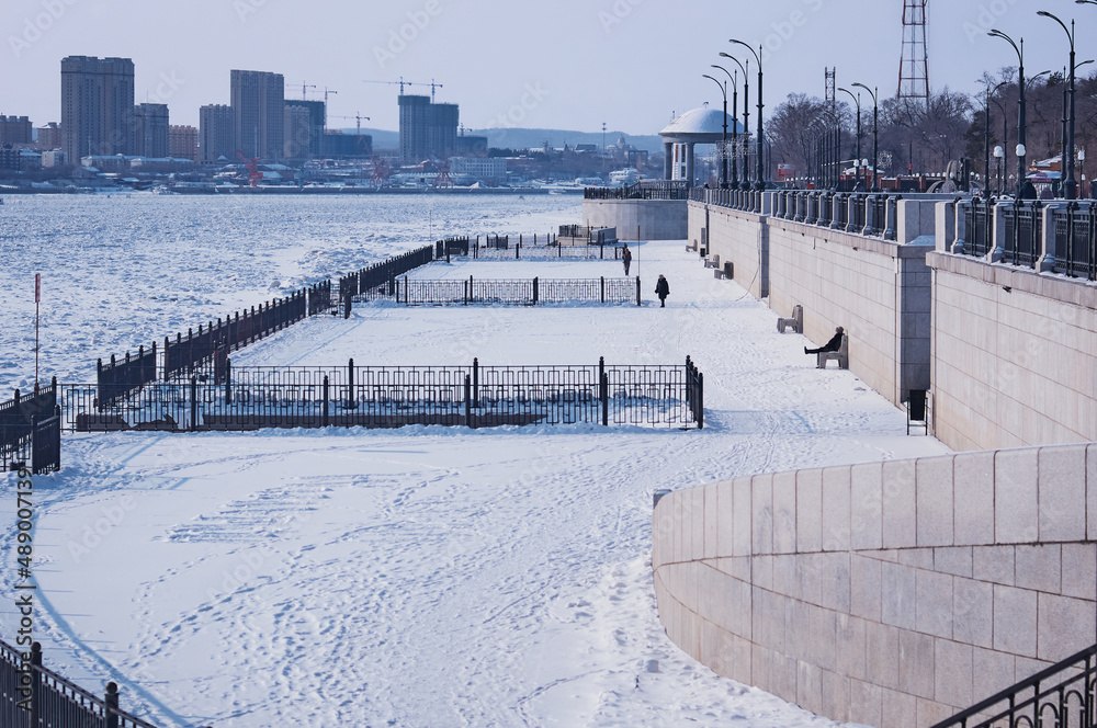 Snow-covered city embankment of the Amur River in winter. The lower level with the townspeople walking along the promenade. Rotunda with a snow-white dome. State border between Russia and China.