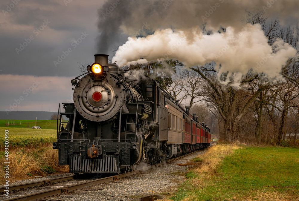 Obraz premium A View of an Antique Freight Steam Train Blowing Smoke Approaching Thru Trees in Late Afternoon on a Cloudy Day