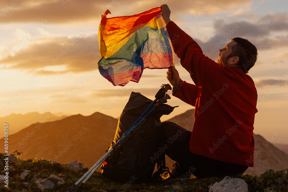 hiker man sitting on the mountain next to his backpack waving a rainbow flag of lgbt pride. sport and inclusion.
