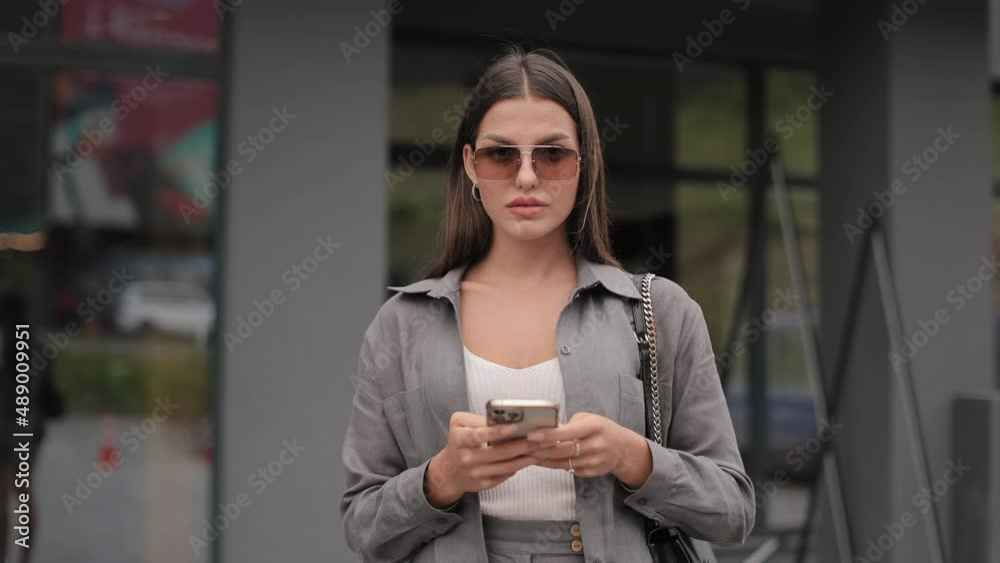 Adorable young woman looking at camera with confidence in eyes while standing on street and holding mobile in hands. Charming lady wearing trendy sunglasses and stylish clothes.