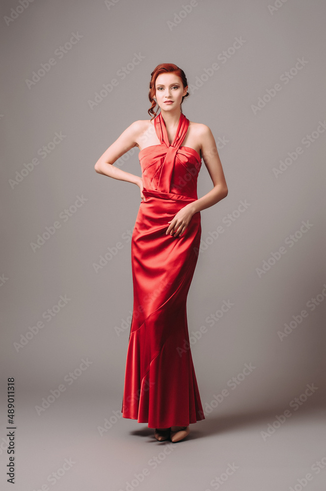 Bright young lady in red long sleeveless dress with gathers and a high halter neckline on grey studio background. Fashionable female look.