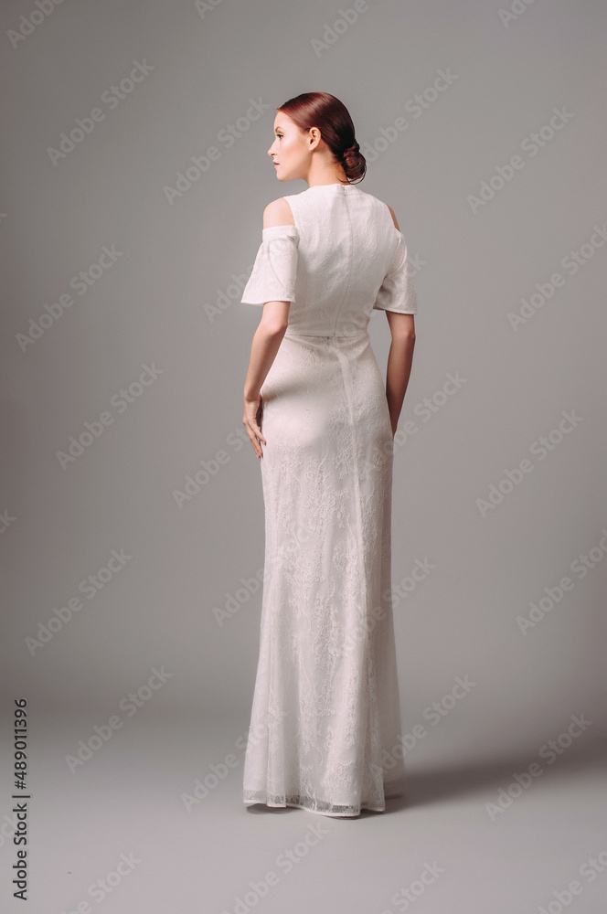Modern bridal look. Happy smiling ginger bride in white wedding gown, rear view. Full-length trumpet dress with floral lace print, off the shoulders sleeves and high neck line. Studio shot. Modern bridal look. Happy smiling ginger bride in white wedding gown, rear view. Full-length trumpet dress with floral lace print, off the shoulders sleeves and high neck line. Studio shot.