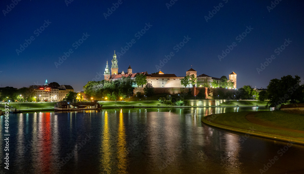 Obraz premium Poland, Krakow. Illuminated royal Wawel castle and cathedral at night and light reflections in Vistula River. Riverside with park, trees, promenade, lanterns, harbor and tourist ship