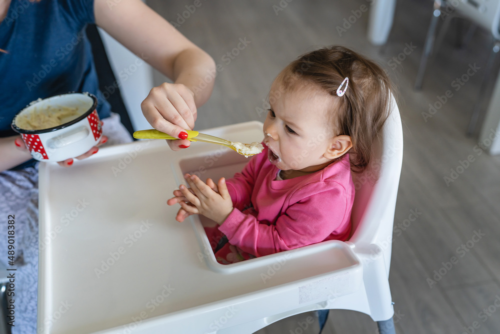One baby small caucasian girl sitting in the feeding high chair while her mother holding spoon with food real people copy space One baby small caucasian girl sitting in the feeding high chair while her mother holding spoon with food real people copy space