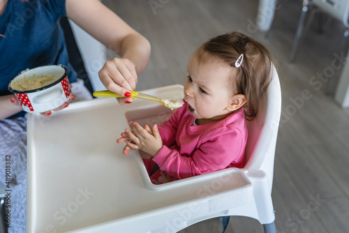 One baby small caucasian girl sitting in the feeding high chair while her mother holding spoon with food real people copy space
