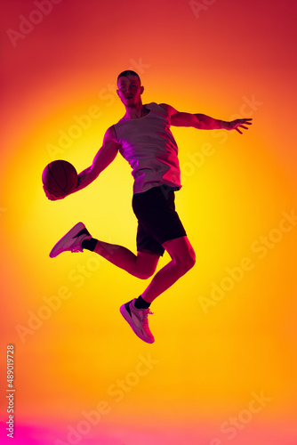 Studio shot of muscled man  basketball player training with ball isolated on gradient yellow purple background in neon light. Beauty  sport  motion  activity concepts.