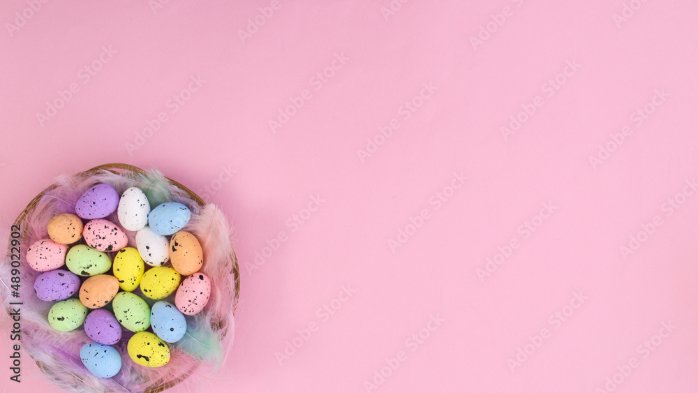 Creative pastel pink copy space background with wooden basket and vibrant colored eggs for Easter holidays. Spring Flat lay concept