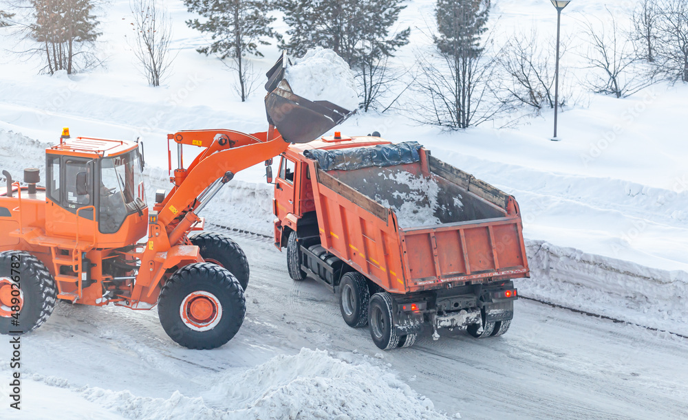 Big orange tractor cleans up snow from the road and loads it into the truck. Cleaning and cleaning of roads in the city from snow in winter. Snow removal after snowfall and blizzards. Big orange tractor cleans up snow from the road and loads it into the truck. Cleaning and cleaning of roads in the city from snow in winter. Snow removal after snowfall and blizzards.