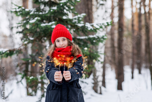 A teenage girl in a red hat and snood holds a sparkler in her hands in a winter forest. Walk in the cold season in warm clothes. Beautiful teenager in nature.