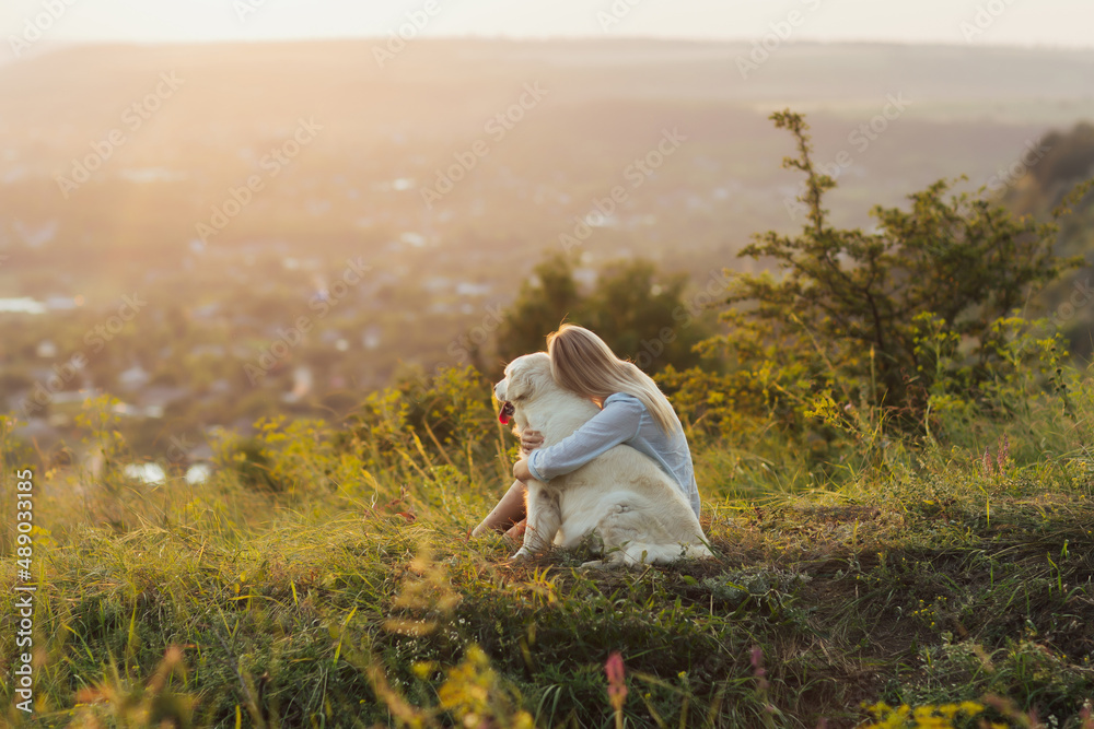Blonde girl hugging a dog in the mountains at sunset. Friendship between woman and dog. Traveling with a pet.