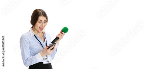 Excited young girl  female journalist holding reporter microphone and looking at phone isolated on white background. Concept of social media  press  news  information