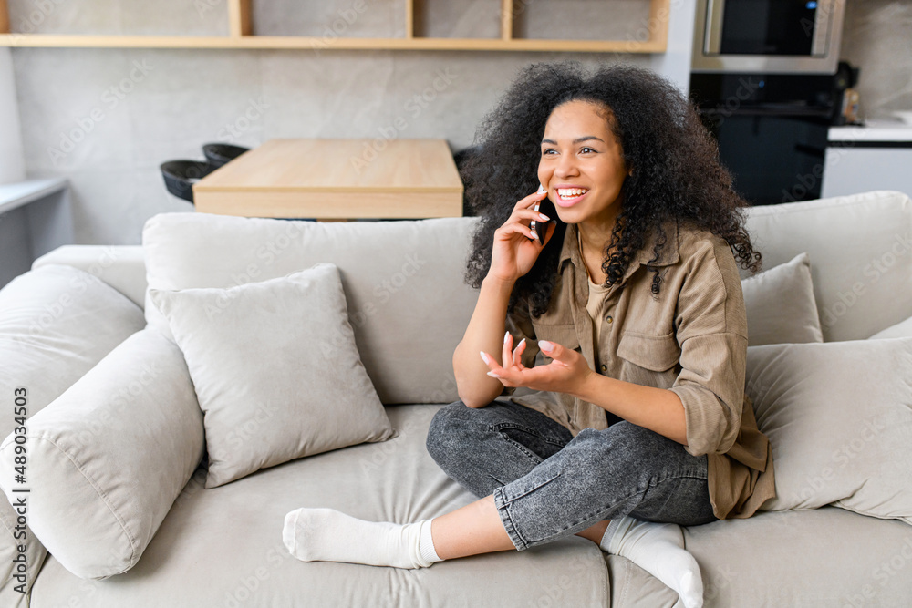 Smiling young multiracial woman talking by smartphone and looks away. Carefree charming lady enjoys phone conversation with friend, sits on sofa, chatting and laughing Smiling young multiracial woman talking by smartphone and looks away. Carefree charming lady enjoys phone conversation with friend, sits on sofa, chatting and laughing
