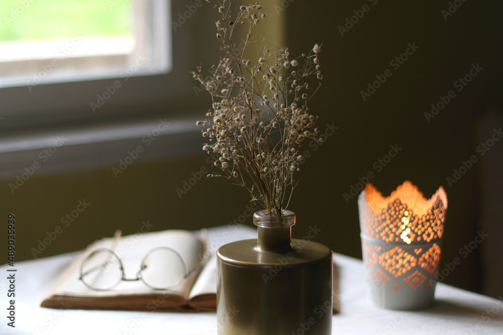 Open book, reading glasses, candle holder with lit candle and vase with gypsophila flowers. Dark background, selective focus. Open book, reading glasses, candle holder with lit candle and vase with gypsophila flowers. Dark background, selective focus.