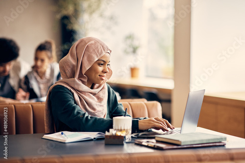 Young smiling Muslim woman e-learning on laptop at cafeteria.