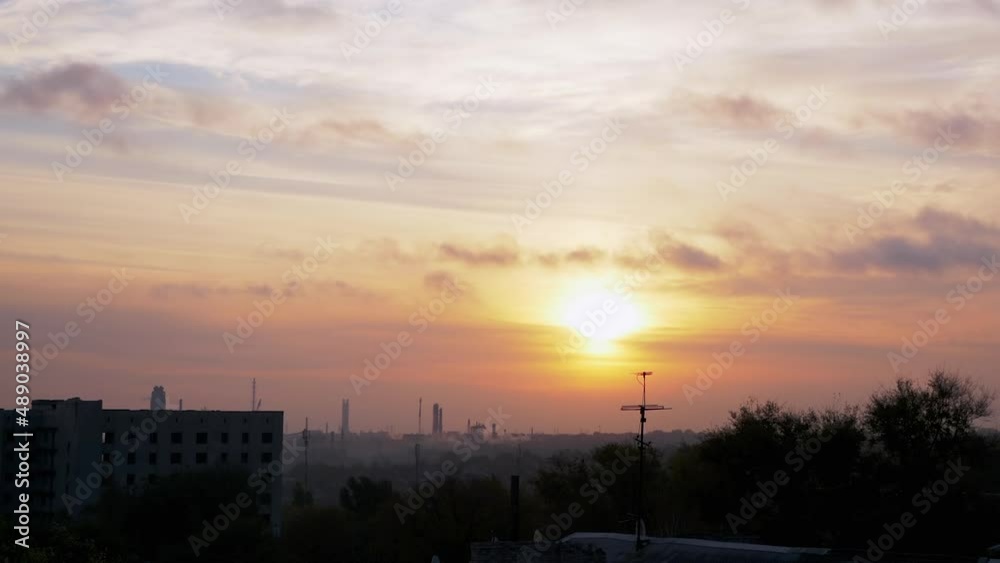 Sunrise Over a Sleeping Industrial City. Disappearing gray smoky clouds move over treetops and unfinished buildings. Rising soft sun over the red-orange horizon. Flying flock of birds. Early morning.