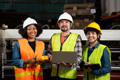 Front view of three factory workers, African woman, Asian man and woman, in a vest and helmet, standing smiling at camera while working with a multimeter, laptop, and tablet held in hand at a factory.