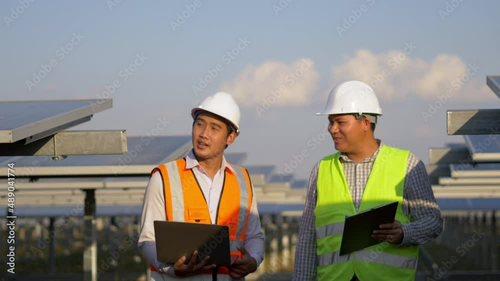 Two Asian young engineers walking along rows of photovoltaic panels in solar farm, They use laptop computer and talking together Two Asian young engineers walking along rows of photovoltaic panels in solar farm, They use laptop computer and talking together