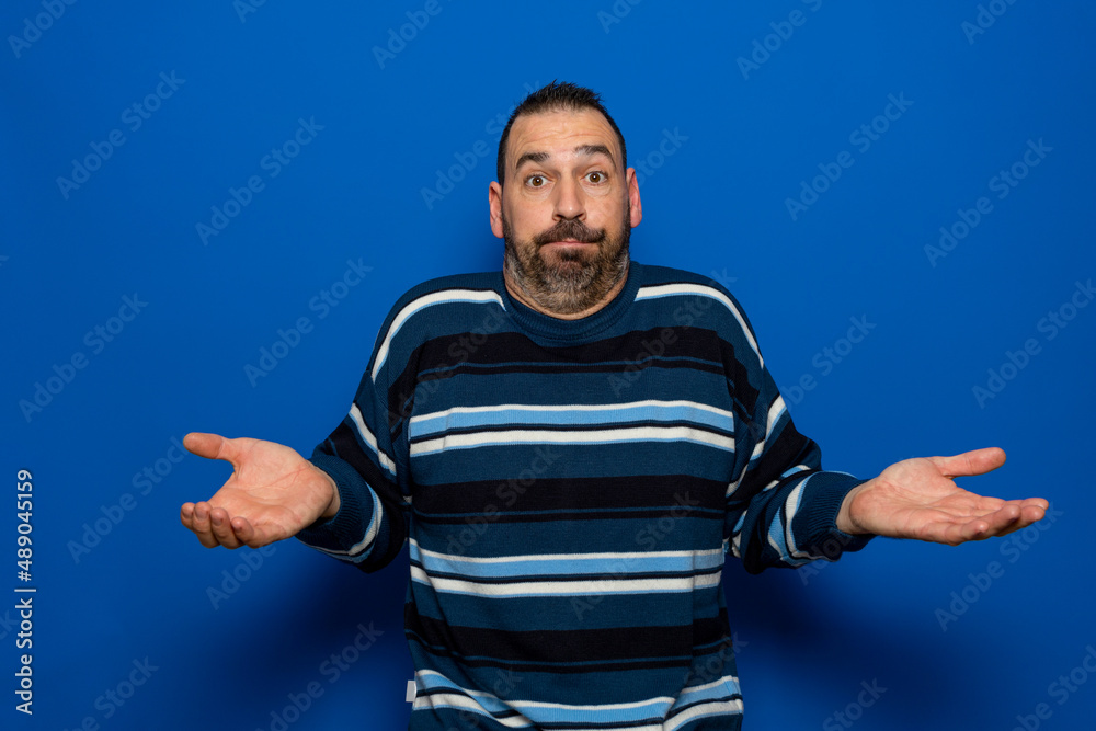 Fototapeta premium Puzzled hesitant bearded fashionable male full of doubts, should decide or choose between two proposals, pouty lips, has indecisive expression, shows uncertainty, gestures against blue background