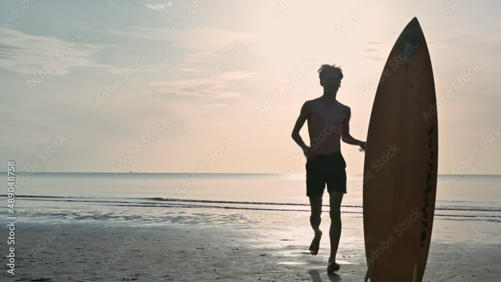 Young asian man running up to picking surfboard on the beach at morning