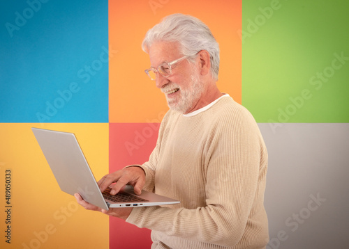 Portrait of adult bearded senior man holding using laptop computer standing over cute colorful background. 70 years old grandfather wearing eyeglasses enjoying technology and social