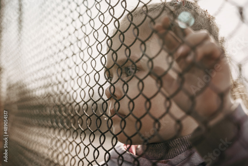 A little refugee girl with a sad look behind a metal fence. The social problem of refugees and internally displaced persons photo