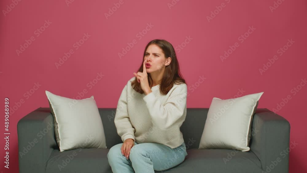 Serious severe beautiful young girl showing silence gesture, ask to stay quiet, keep secret, sitting on sofa Serious severe beautiful young girl showing silence gesture, ask to stay quiet, keep secret, sitting on sofa