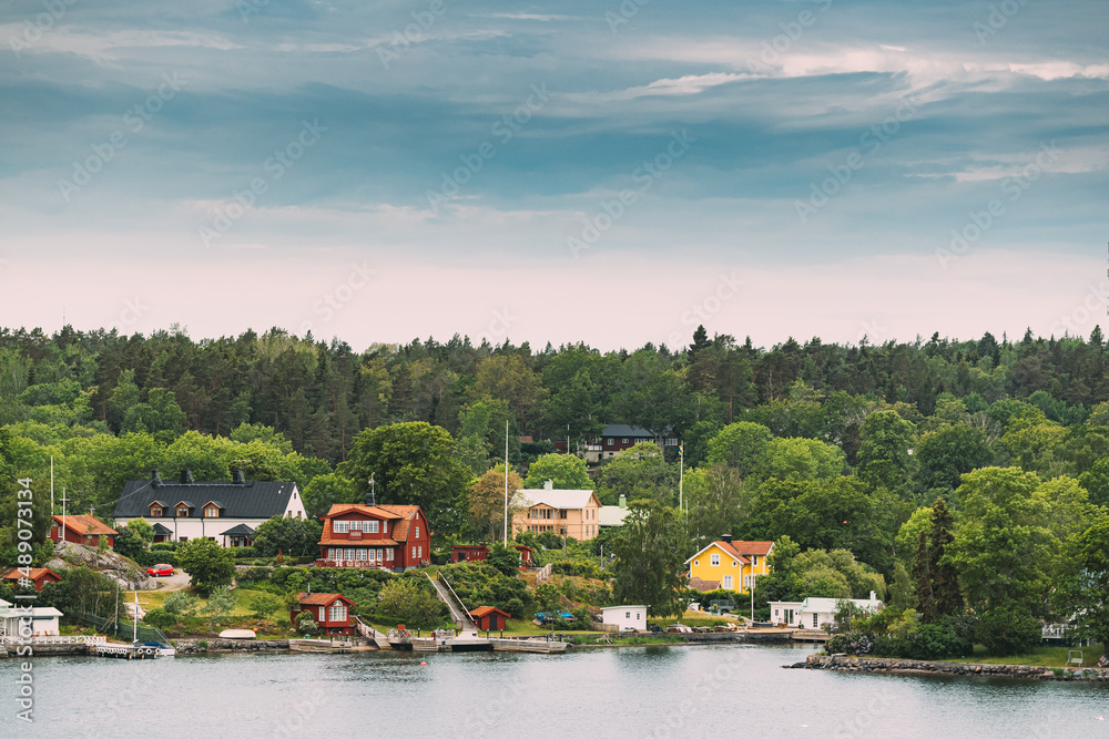 Sweden. Many Beautiful Red Swedish Wooden Log Cabins Houses On Rocky Island Coast In Summer Sunny Evening. Lake Or River Landscape.