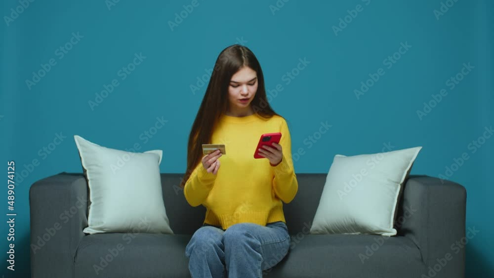 Smiling young girl holding bank credit card, phone sitting on couch, making purchase in online store. E-commerce Smiling young girl holding bank credit card, phone sitting on couch, making purchase in online store. E-commerce