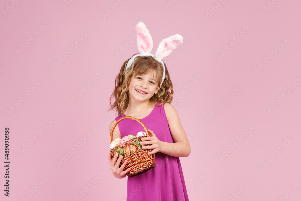 Little beautiful smiling girl in Easter bunny ears headband holds in hands a basket with Easter eggs on pink pastel background Little beautiful smiling girl in Easter bunny ears headband holds in hands a basket with Easter eggs on pink pastel background
