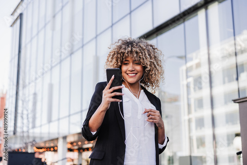 beautiful smiling business woman using mobile phone and headphones in city. Buildings background