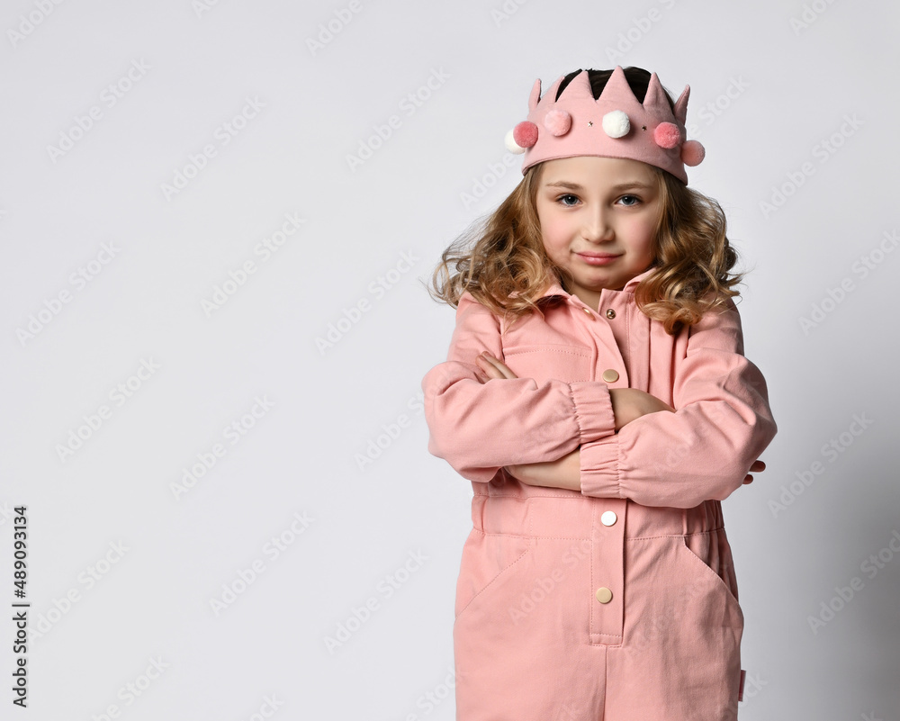 Portrait of a cute girl with a plush crown, stands with her arms crossed over her chest, in a closed pose. in a dusty rose cotton summer jumpsuit. Portrait of a cute girl with a plush crown, stands with her arms crossed over her chest, in a closed pose. in a dusty rose cotton summer jumpsuit.