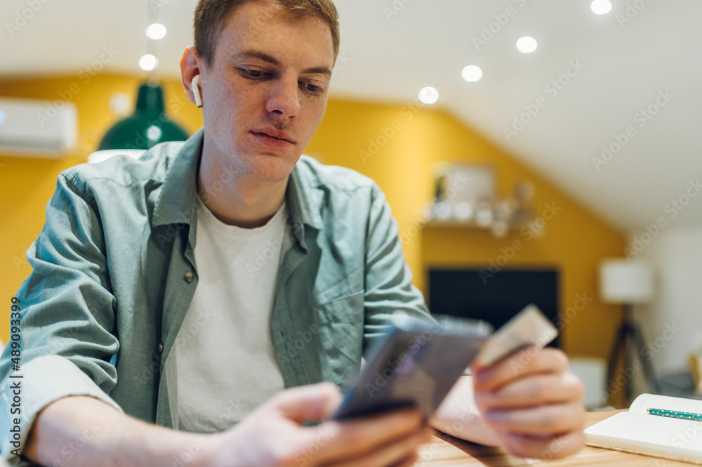 Fototapeta premium Redhead man using smartphone and a credit card in the kitchen at home