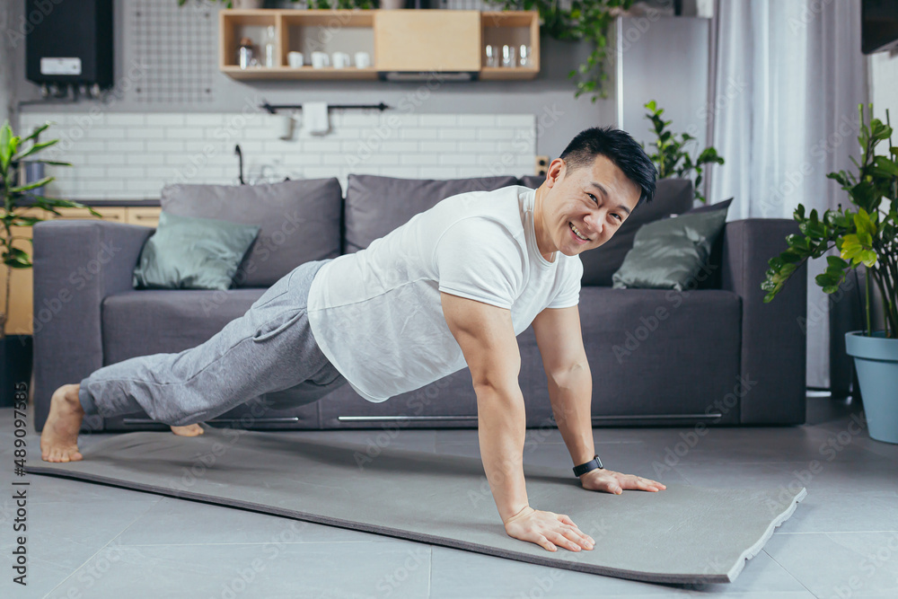 Portrait of an Asian man at home, doing fitness and sports, at home in the living room, on a sports mat, pushing off the floor smiling and looking at the camera Portrait of an Asian man at home, doing fitness and sports, at home in the living room, on a sports mat, pushing off the floor smiling and looking at the camera