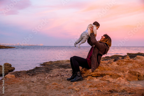 Lifestyle of a family on the beach, a baby having fun and lifting him up with his mother sitting by the sea at sunset