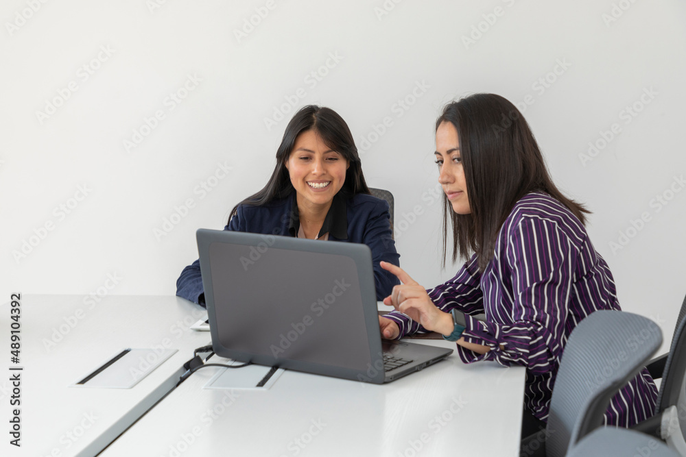 Two female co-workers chat while reviewing a project on their laptop. Latin women working in office Two female co-workers chat while reviewing a project on their laptop. Latin women working in office