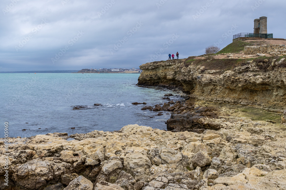 View of the rocky coast of the Black Sea. People are walking along the shore. On the hill is an old signal bell. Cloudy windy weather. Chersonesus, Sevastopol, Crimea.