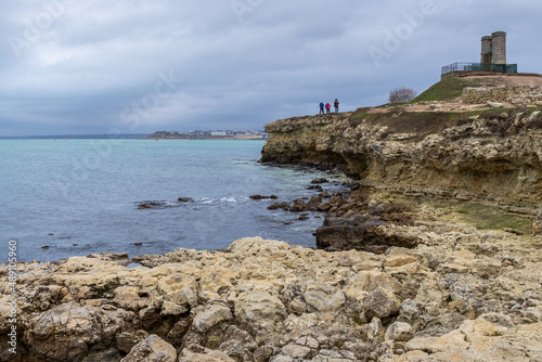 View of the rocky coast of the Black Sea. People are walking along the shore. On the hill is an old signal bell. Cloudy windy weather. Chersonesus, Sevastopol, Crimea. © Andrei Stepanov