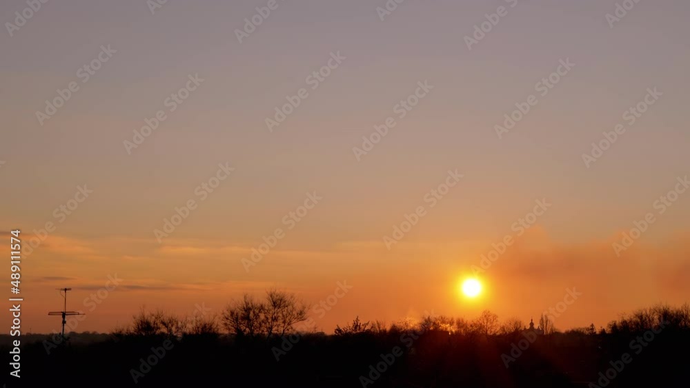 Sunrise through the Clouds over the Treetops and the Dark Horizon. Windy weather, moving clouds, fog, smog. The bright orange sun slowly rises in the sky. Time lapse. Sunrise through the Clouds over the Treetops and the Dark Horizon. Windy weather, moving clouds, fog, smog. The bright orange sun slowly rises in the sky. Time lapse.