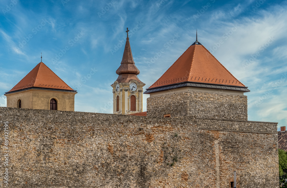 The towers of Varpalota Hungary. Two square shape medieval castle tower of the Thury castle with red roof and the baroque clock tower of the municipal catholic church